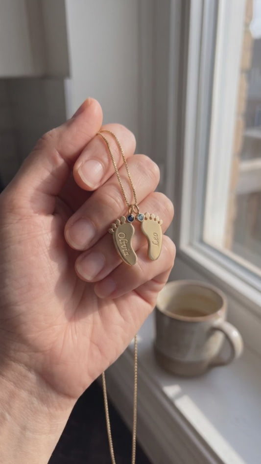 A hand holds the Little Feet Birthstone Necklace at a sunny kitchen window
