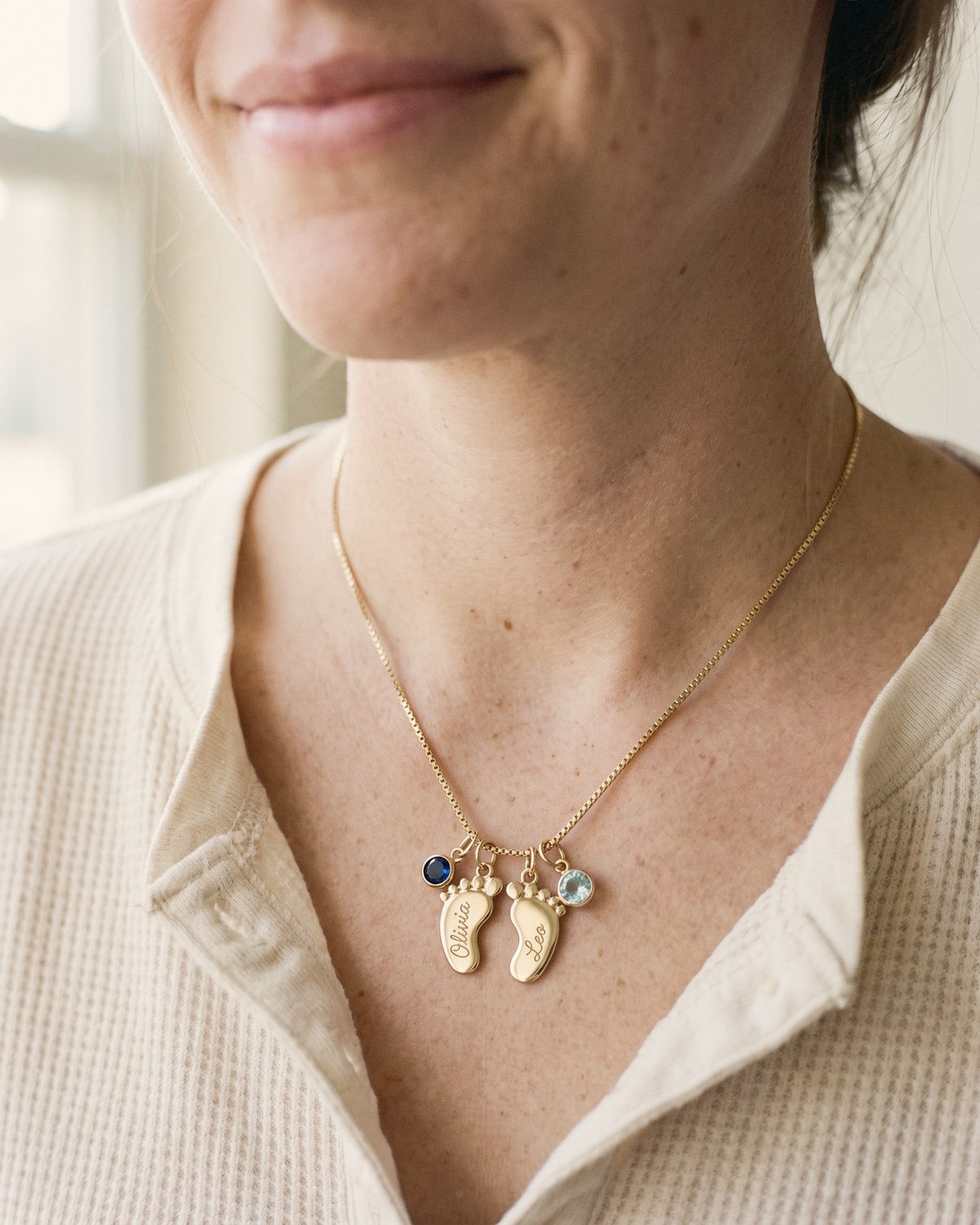 A mom wearing two gold baby-feet charms engraved Olivia and Leo with birth-month stones
