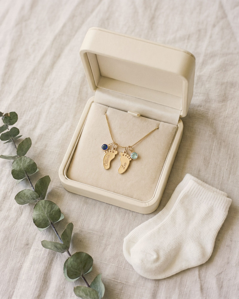The baby-feet necklace in its gift box beside tiny knit socks