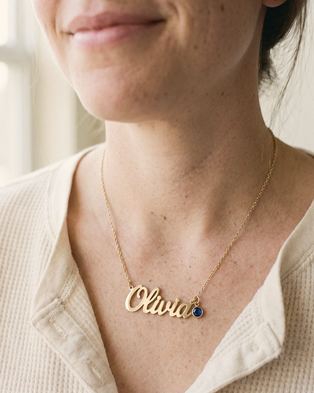 A new mom wearing a gold name necklace reading Olivia with a small blue birthstone